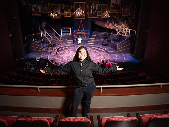 A person stands smiling in the seating area of a theater, looking toward the camera, with an elaborate stage set in the background.