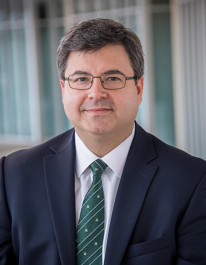 A man in a suit and tie is posing for a formal portrait, wearing glasses and standing in front of a blurred backdrop.