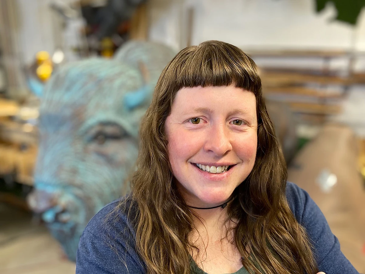 A person with long hair is smiling in the foreground, with a large sculpture of a blue buffalo in the background.