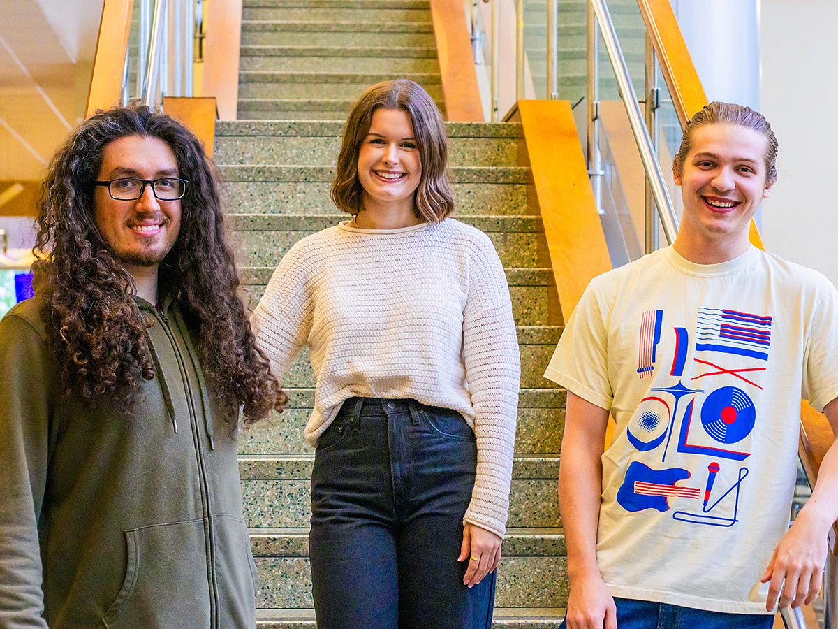 Three people are smiling while standing on a staircase indoors.